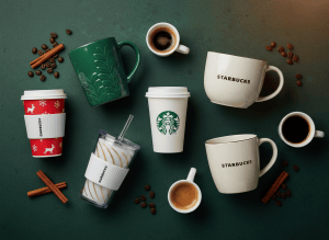 A beautiful flat lay of Starbucks cups and mugs on a dark green background, including iconic green Starbucks logo cups, ceramic mugs, and holiday cups arranged aesthetically. Warm lighting, clean and modern commercial photography style.